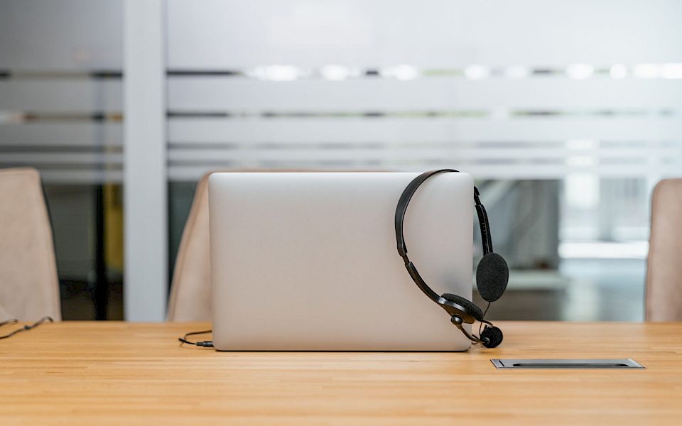 Headset hängt über einem silbernen Laptop auf einem Holztisch in einem hellen Büro